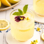A glass of lemonade with lemon, mint, and edible flowers on a white coaster, surrounded by lemon halves and flowers, evokes the refreshing aesthetic of a whiskey sour.