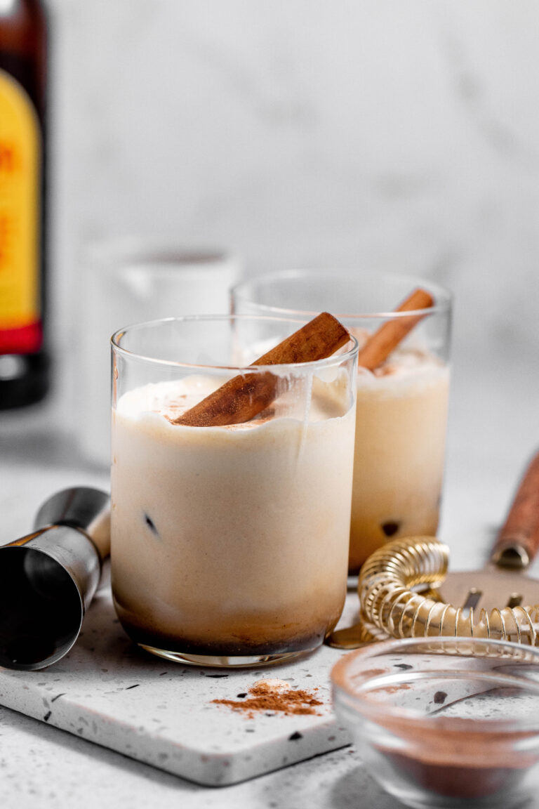 Two creamy whiskey sours with ice and cinnamon sticks are in the foreground, with a bottle, jigger, and cocktail strainer in the background.