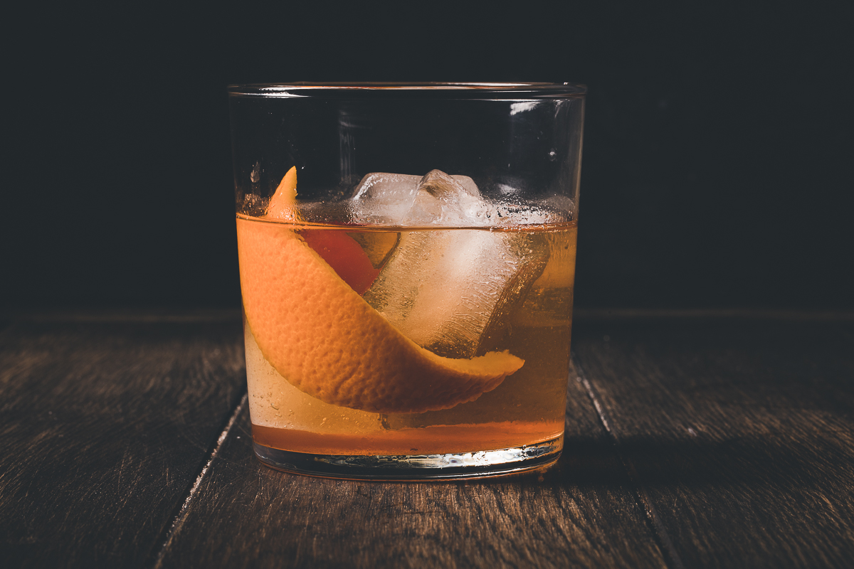 An amber whiskey sour with an orange peel and ice cubes rests on a wooden surface against a dark background.