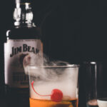 A smoked bourbon with orange and cherry garnishes evokes a whiskey sour, with a bottle of Jim Beam Kentucky Bourbon Whiskey standing proudly behind.