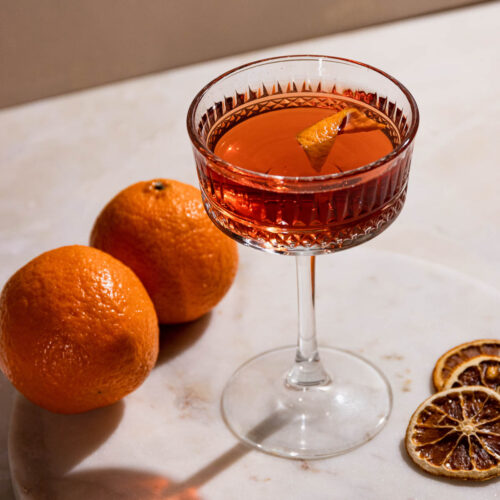 An el presidente cocktail with an orange twist in a cocktail glass on a white table casting a long shadow.