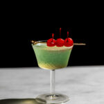 Last word cocktail in a glass with a cocktail pick garnished with three cherries.