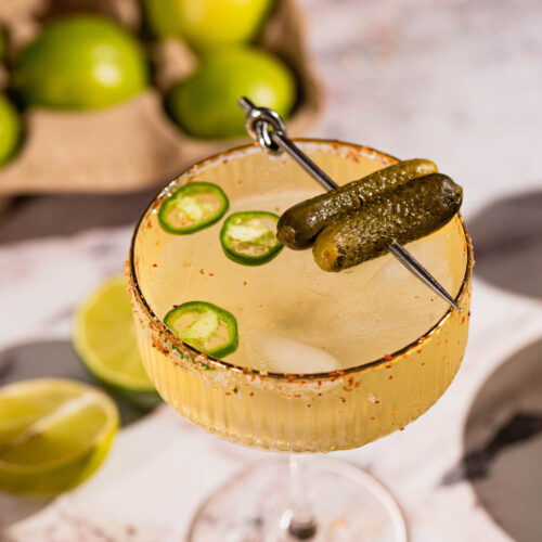 Pickle margarita in a cocktail glass garnished with jalapenos and pickles.