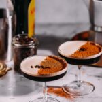 Two chai espresso martinis with cocoa powder rest on a marble surface surrounded by scattered coffee beans, a liqueur bottle, a jar, and an espresso maker.