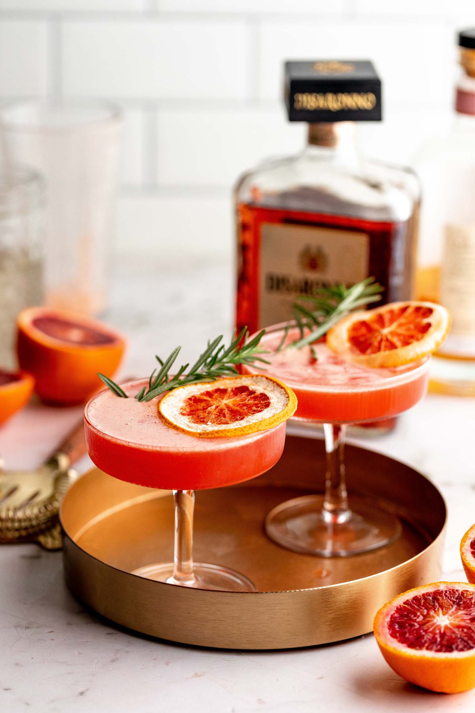 Two amaretto sour cocktails on a tray, garnished with blood orange slices and rosemary, with bottles and halved blood oranges in the background.