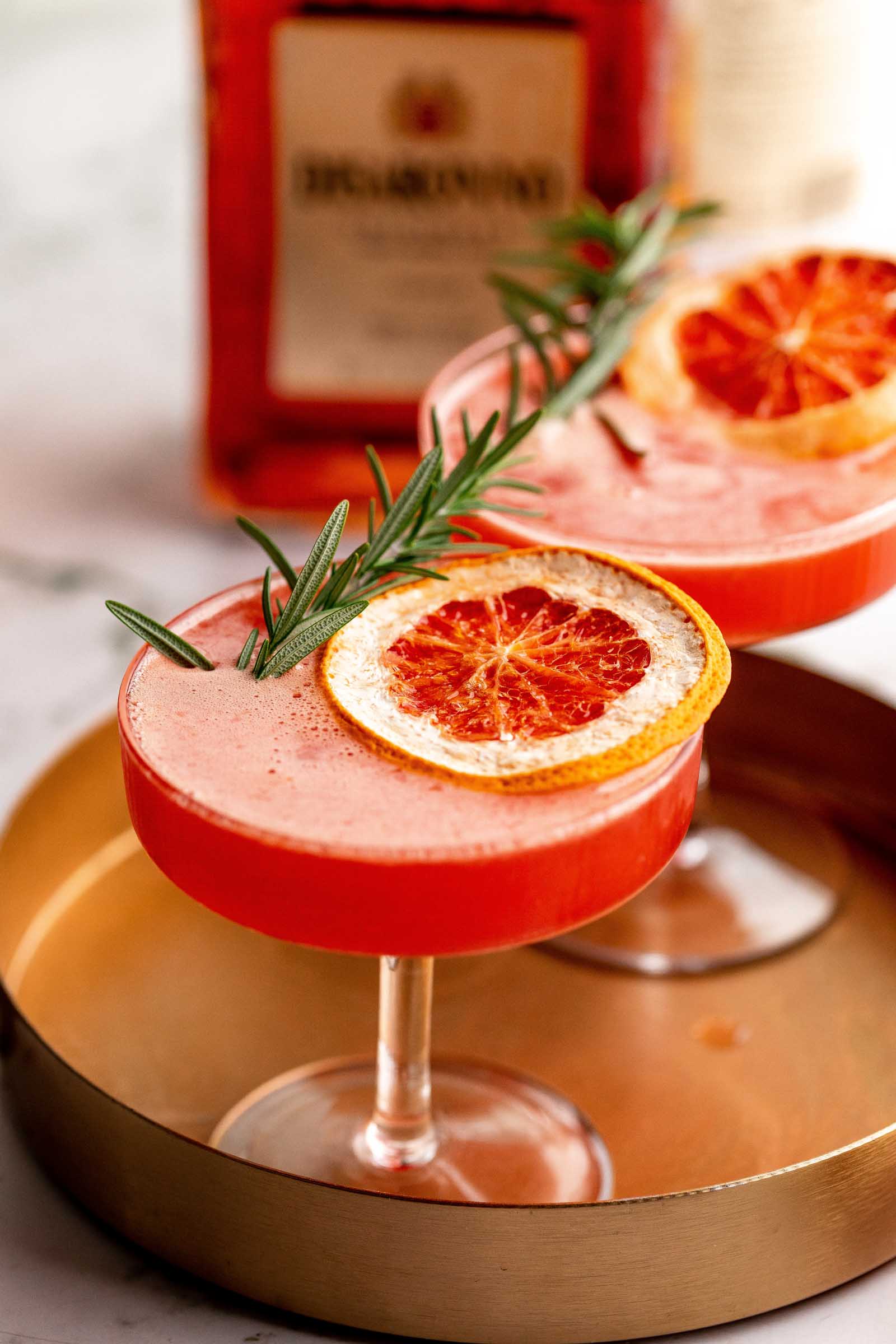 A gold tray holds two amaretto sour cocktails garnished with dried orange slices and rosemary, with a bottle in the background.