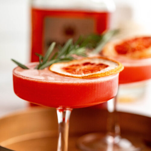 An amaretto sour cocktail in a coupe glass, garnished with dried blood orange and rosemary, served on a brass tray.