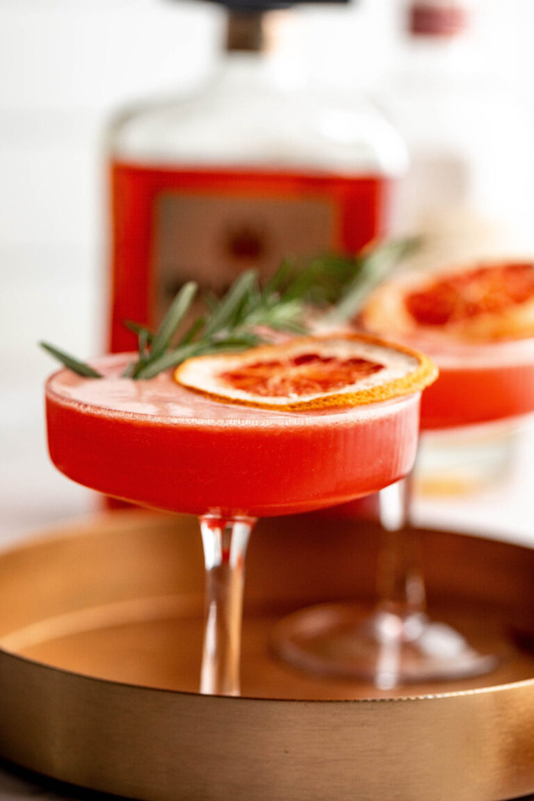 An amaretto sour cocktail in a coupe glass, garnished with dried blood orange and rosemary, served on a brass tray.