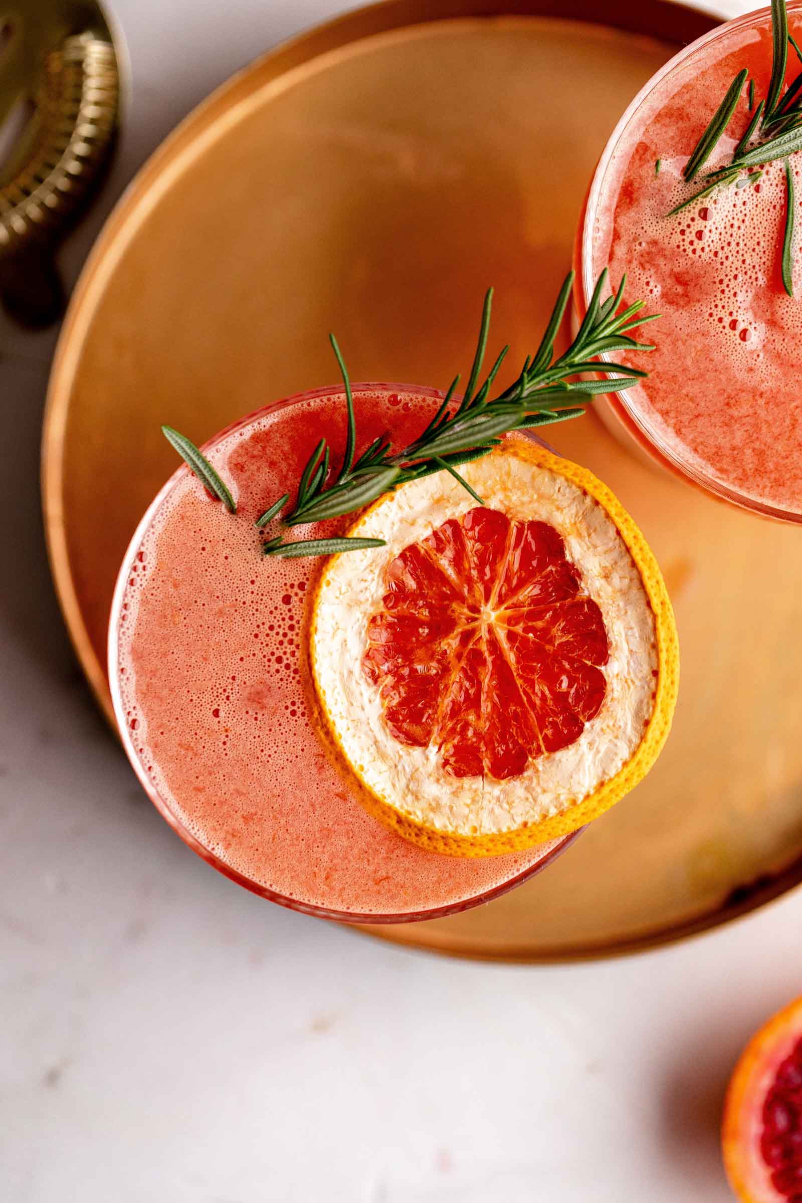 An amaretto sour cocktail with a grapefruit slice and rosemary sprig garnish is served in a glass on a copper tray.