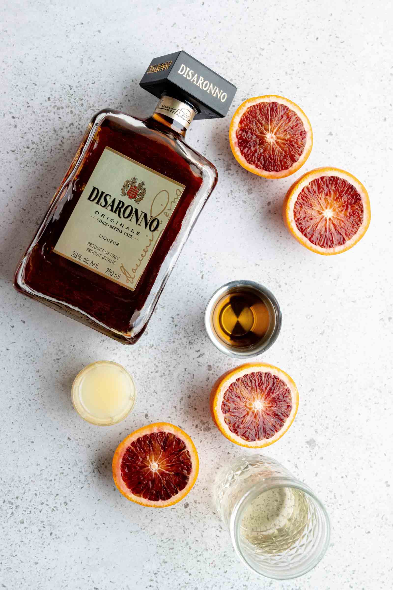 A bottle of Disaronno liqueur, sliced blood oranges, and small cups of liquid are arranged on a speckled surface.