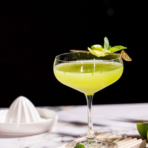 A basil martini garnished with lime twist and mint sits on a marble surface with a citrus juicer in the background.