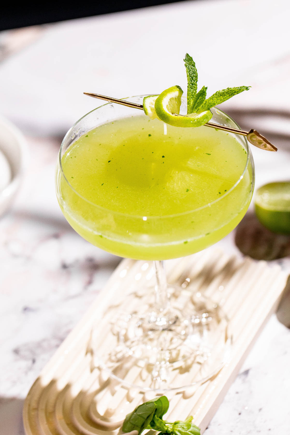 A basil martini cocktail in a coupe glass, garnished with lime twist and mint leaves, rests on a light tray.