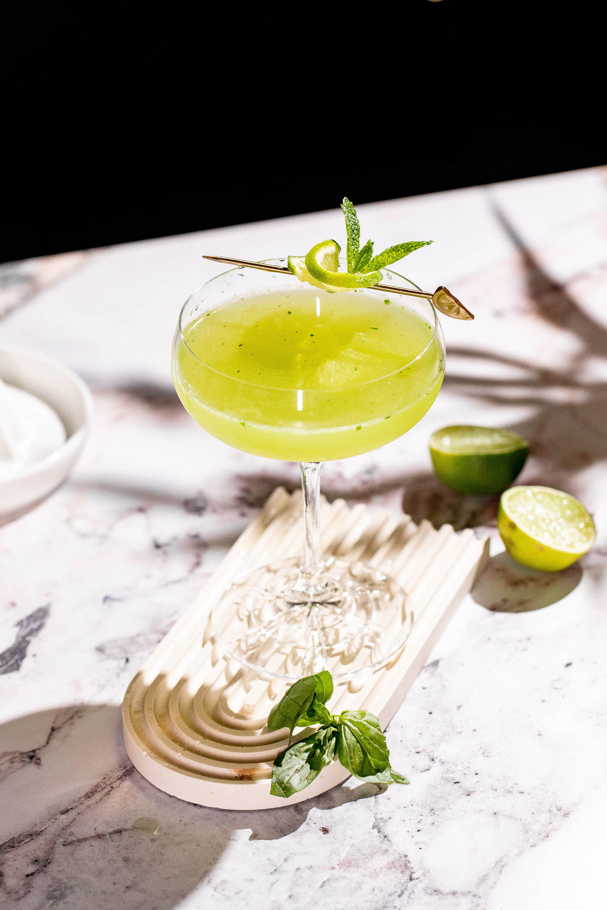 A green basil martini garnished with lime and mint sits on a patterned tray on a marble surface, surrounded by lime halves and a mint sprig.