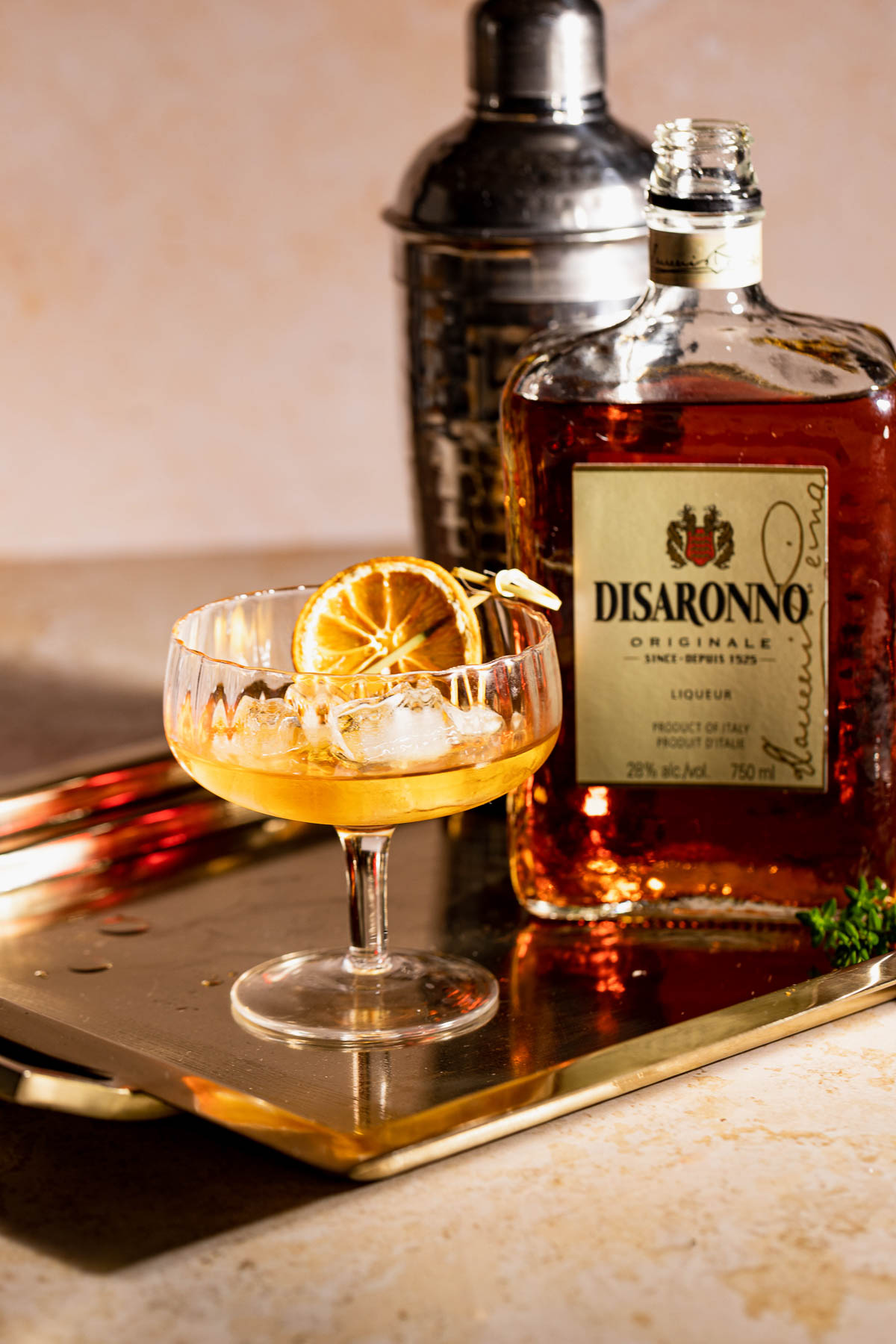 A French Connection cocktail with ice and a dried orange slice sits on a metallic tray beside a bottle of Disaronno liqueur and a shaker.