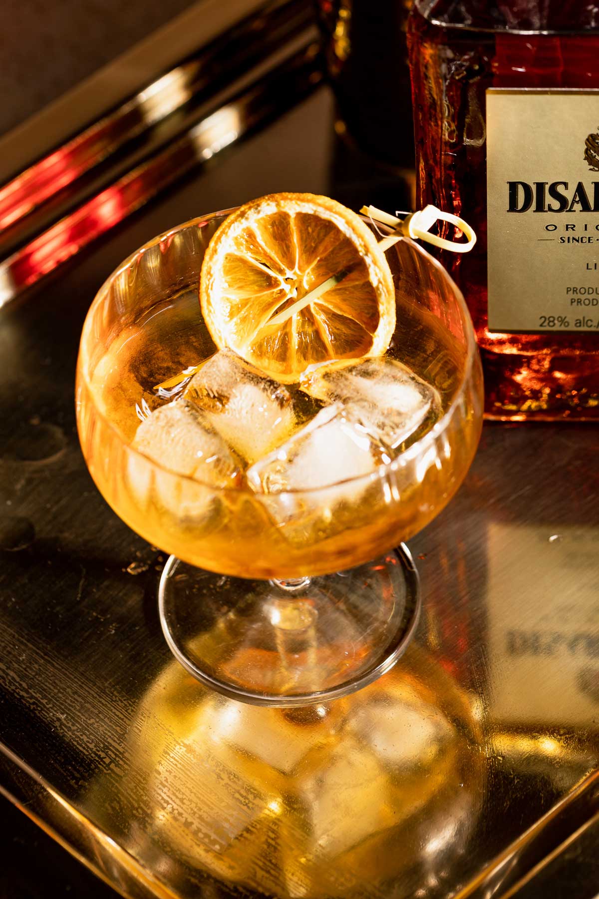 A French connection cocktail with ice and a dried orange slice sits on a reflective surface beside a bottle of Disaronno.