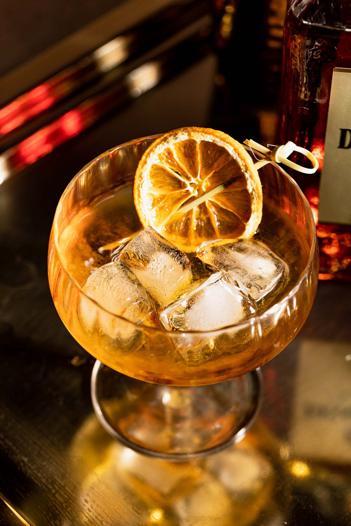 A French connection on ice with a dried lemon slice sits on a reflective gold surface.