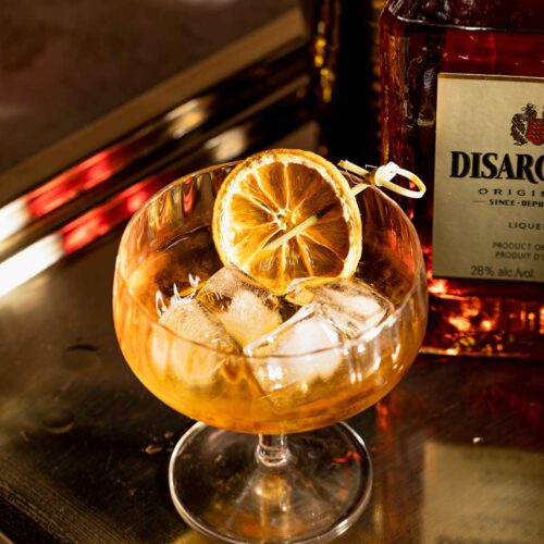 A French Connection cocktail with ice and dried citrus is served in a glass on a gold tray, accompanied by a bottle of Disaronno.