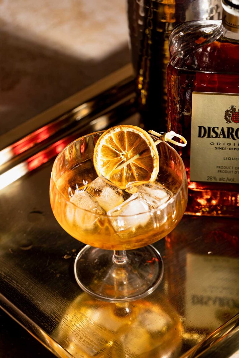 A French Connection cocktail with ice and dried citrus is served in a glass on a gold tray, accompanied by a bottle of Disaronno.