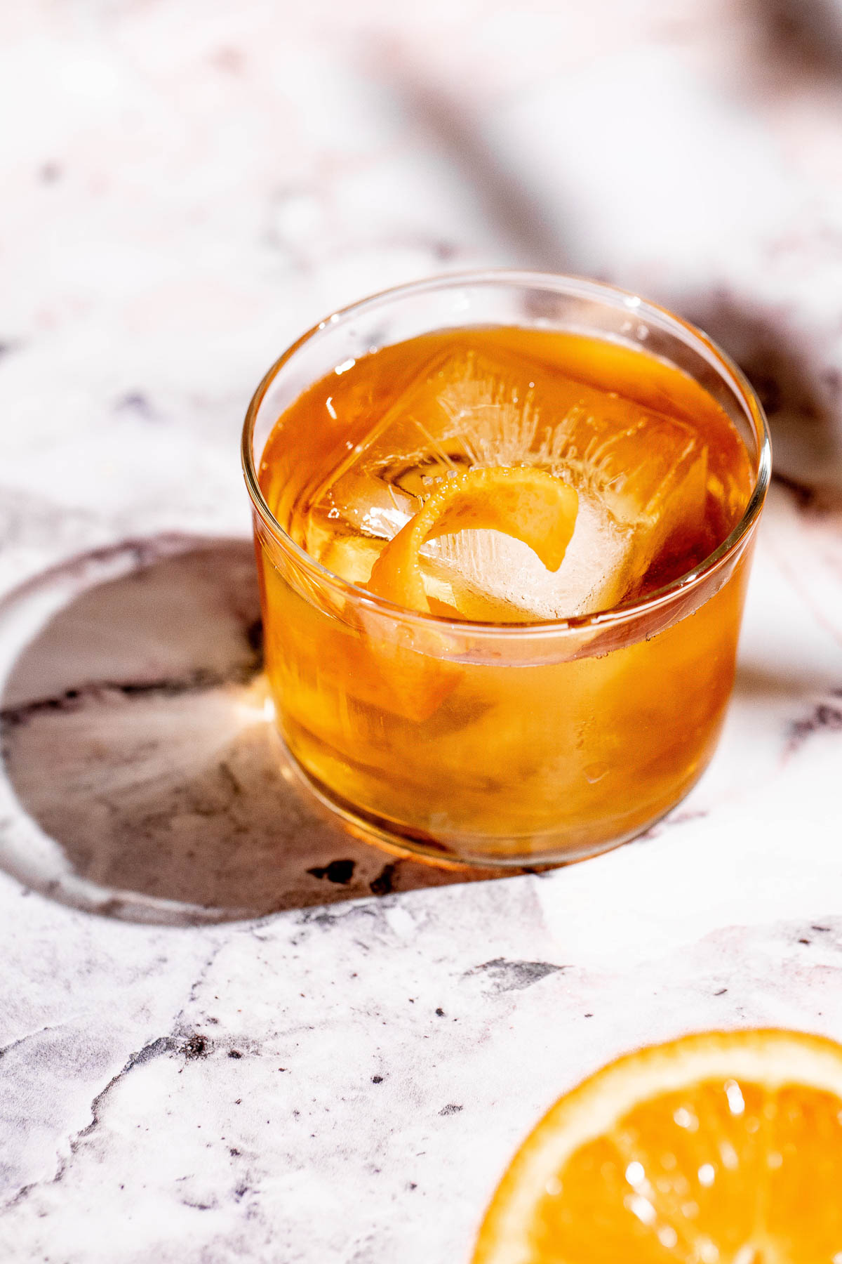 An amber godfather cocktail with ice and an orange peel garnish sits on a marble surface next to an orange slice.