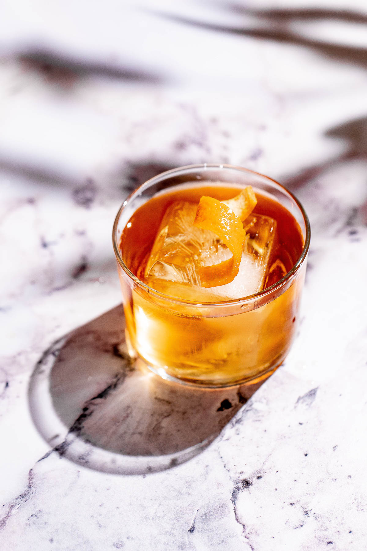 A godfather cocktail with ice and an orange twist casts a shadow on marble.