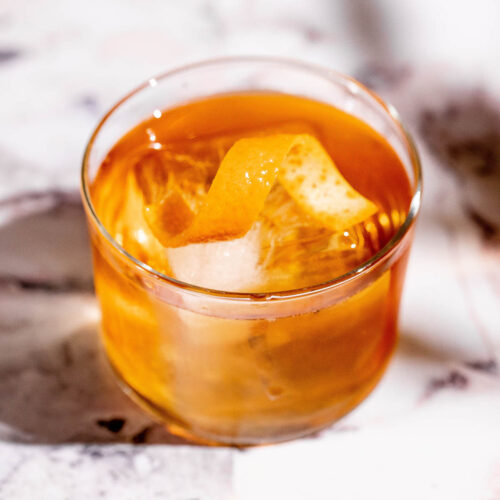 A godfather cocktail with ice and an orange peel garnish sits on a light marbled surface.