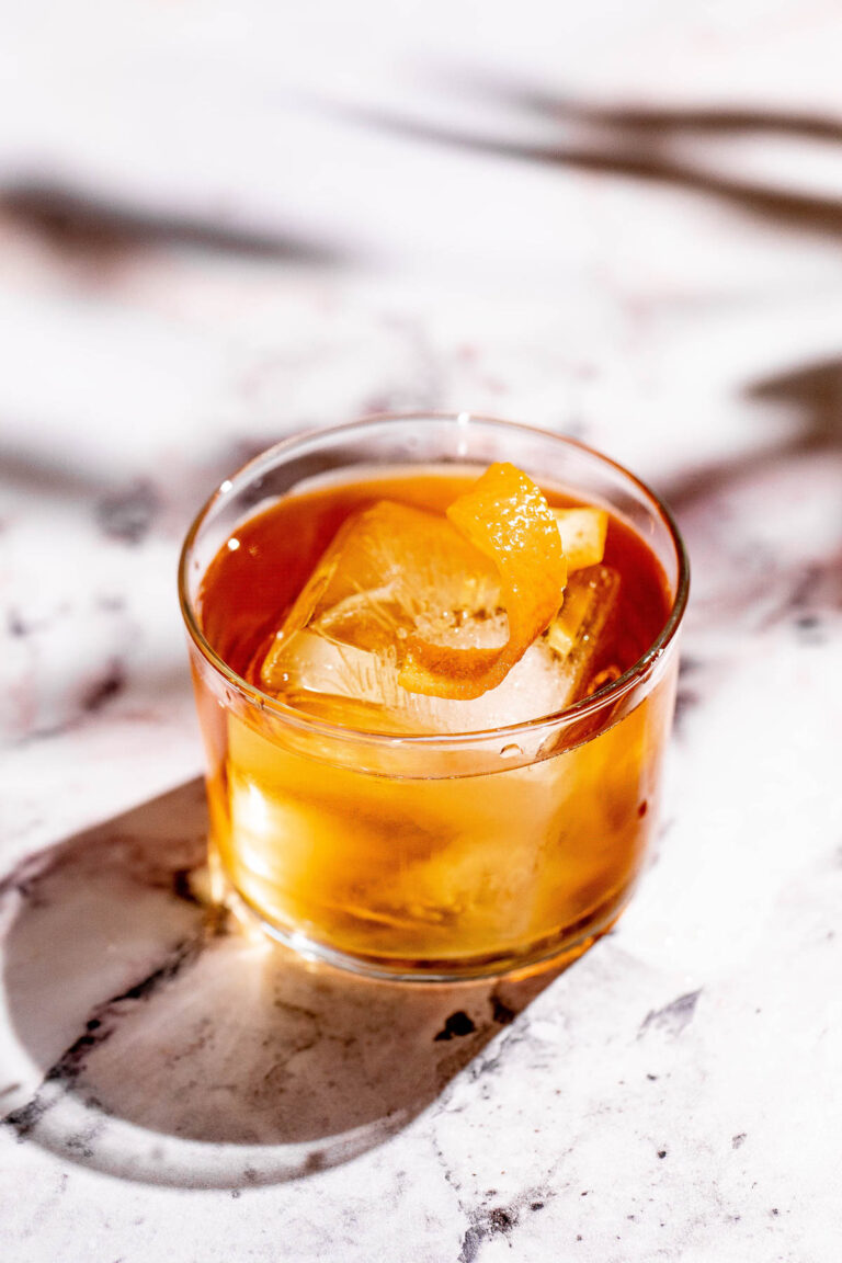 An amber godfather cocktail with an orange peel garnish and large ice cubes sits on a white marble surface.