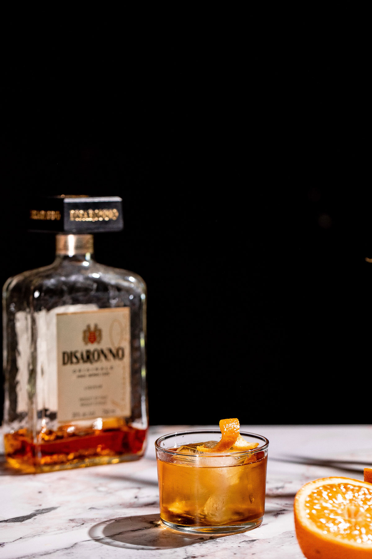 A godfather cocktail with an orange twist is on a marble surface, with a bottle of Disaronno and a sliced orange in the background against a dark backdrop.