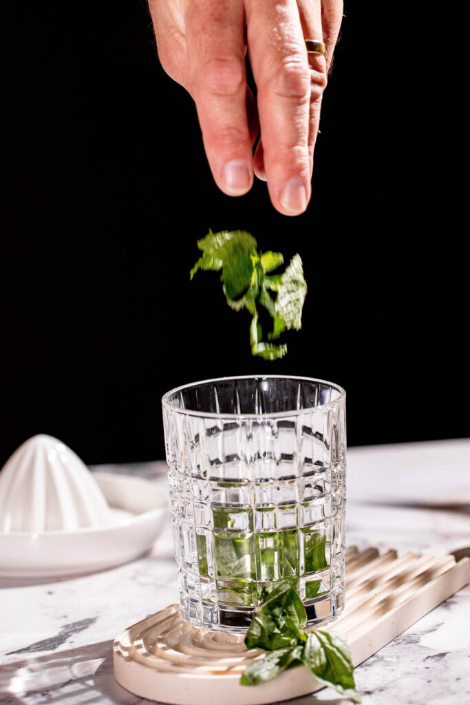 A hand drops basil leaves into a patterned glass on a white coaster, with a lemon squeezer in the background.