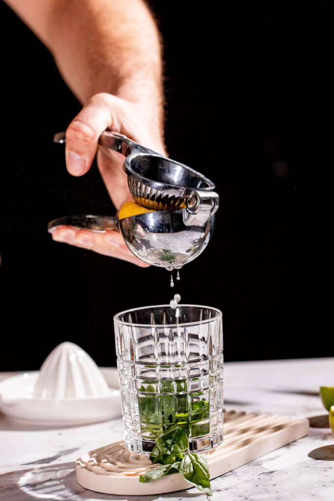 Using a metal juicer, a lemon slice is squeezed over herb-filled glass on marble.