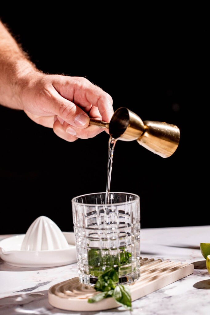 A hand pours simple syrup from a brass jigger into a glass with basil leaves on a marble surface, surrounded by a citrus juicer and lemon slices.