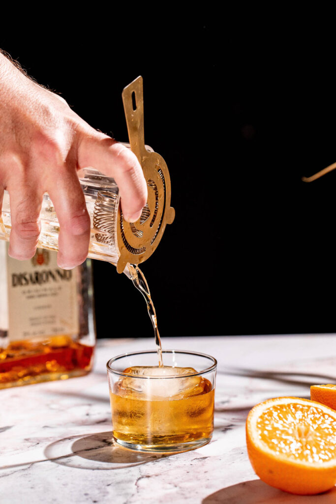 A godfather cocktail is poured through a strainer into an ice-filled glass on a marble surface, with a bottle and halved orange in the background.