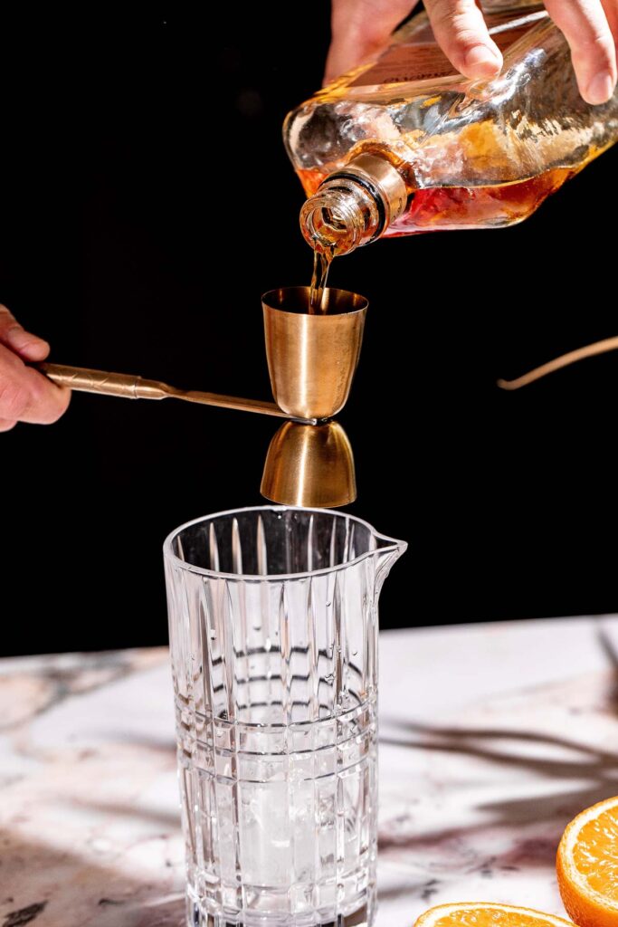 A liquid is poured from a bottle into a jigger above a glass, with citrus slices on the marble surface nearby.