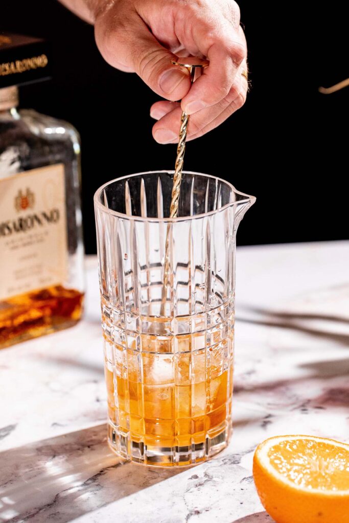 A twisted metal stirrer mixes an amber liquid with ice in a glass pitcher, next to a halved orange and a labeled bottle.