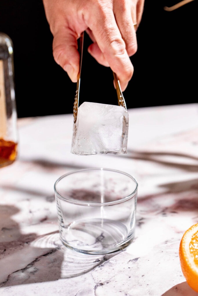 A hand uses tongs to place an ice cube into an empty glass on a marble surface near a sliced orange.