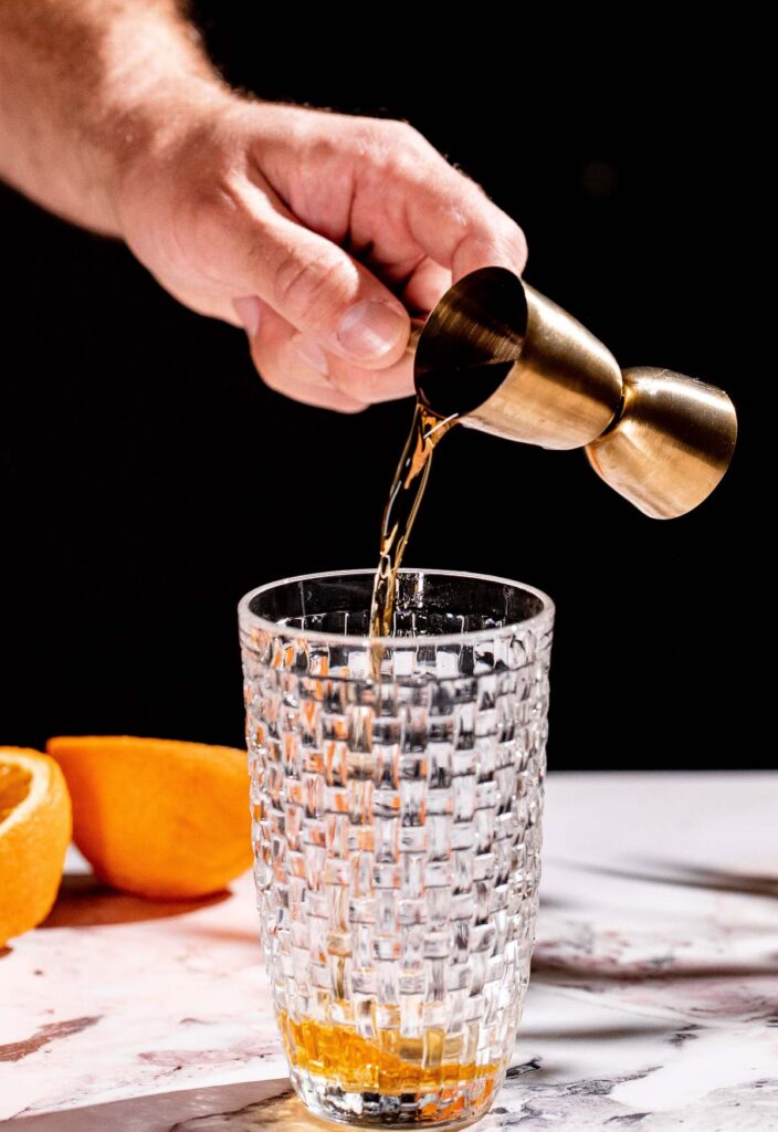 Pouring amaretto from a brass jigger into a textured glass, with an orange half in the background.