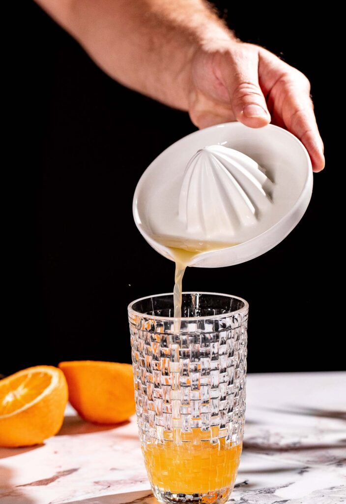 Freshly squeezed orange juice is poured from a ceramic juicer into a textured glass, with halved oranges on the table.