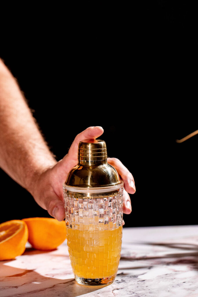 A person holds a gold-topped, textured cocktail shaker with orange liquid on a marble surface next to orange halves.