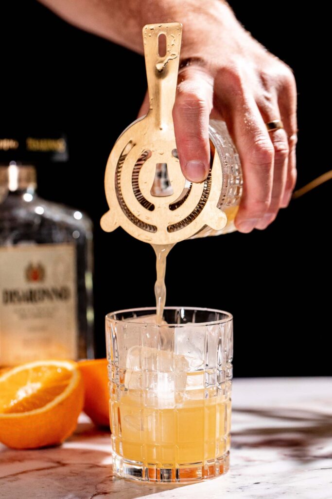 A hand pours an amaretto stone sour cocktail through a strainer into an ice-filled glass, surrounded by a bottle and sliced orange.