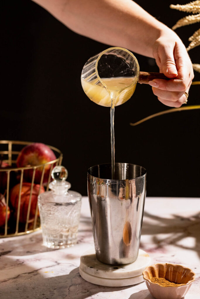 A hand pours apple cider from a glass cup into a metal shaker on a marble counter, crafting an Apple Cider Margarita with apples and a glass container in the background.