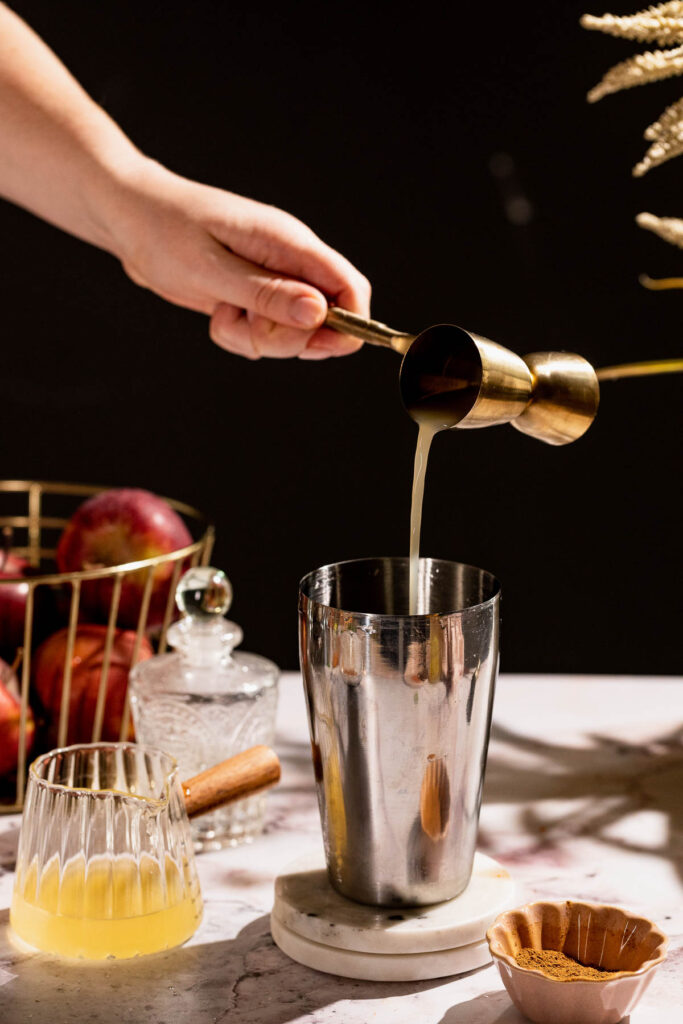 A hand pours liquid from a jigger into a cocktail shaker on a marble counter to craft an Apple Cider Margarita, surrounded by a glass, bottle, and rustic basket of apples.