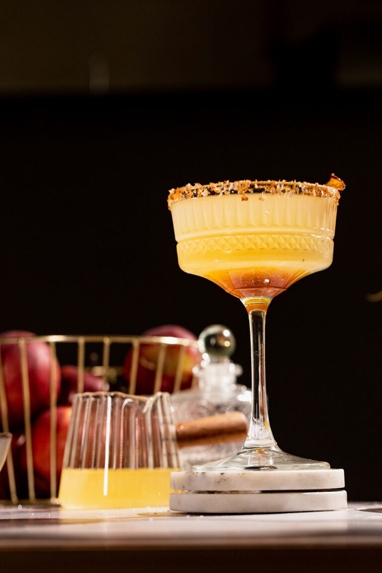 A textured glass with a sugar-rimmed fancy Apple Cider Margarita sits on a coaster, accompanied by apples in a wire basket and a glass of light yellow liquid.