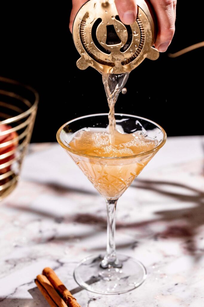 A light brown liquid resembling an apple cider martini is strained from a cocktail shaker into a martini glass on a marble surface, with two cinnamon sticks beside it.