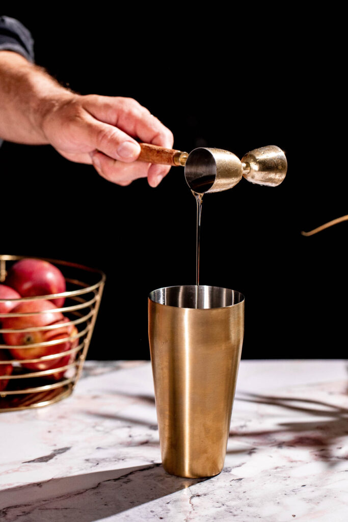 A hand pours liquid from a jigger into a metal shaker on a marble surface, while red apples in a basket add vibrancy to the scene of an apple cider martini in progress.