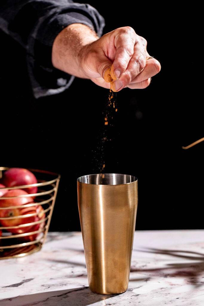 A hand sprinkles spice into a gold shaker on a marble countertop to craft the perfect apple cider martini, with apples in a wire basket echoing the drink's flavors.