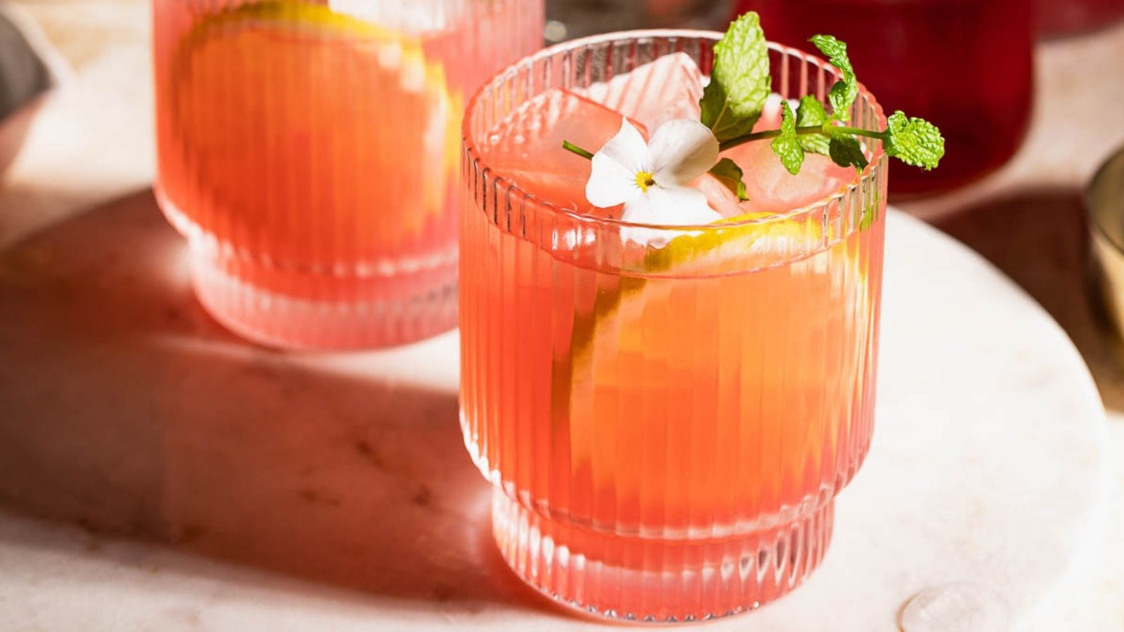 Two pink cocktails in ribbed glasses, garnished with lemon slices, mint, and a white flower, sit on a round marble surface.