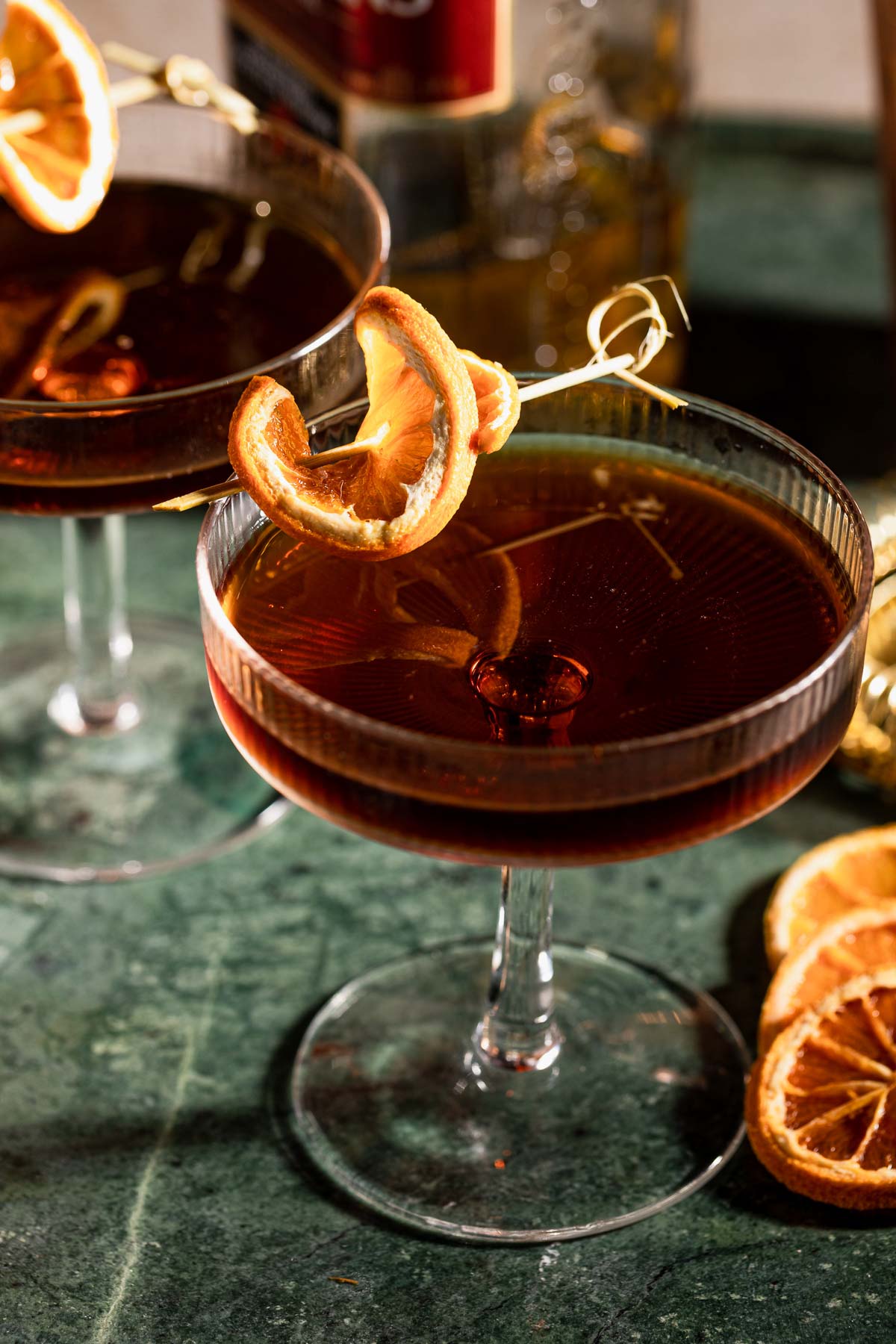 Two coupe glasses containing revolver cocktails, each garnished with a dried orange slice on a skewer, rest on a green marble surface.