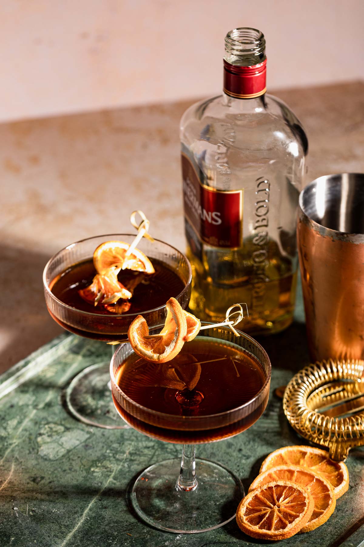 Two cocktail glasses filled with a dark revolver drinks garnished with dried orange slices sit on a table alongside a bottle, shaker, and additional garnishes.