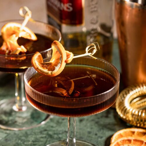 Two dark revolver cocktails with dried orange slices sit on a green table, accompanied by a bottle, shaker, and additional orange slices in the background.