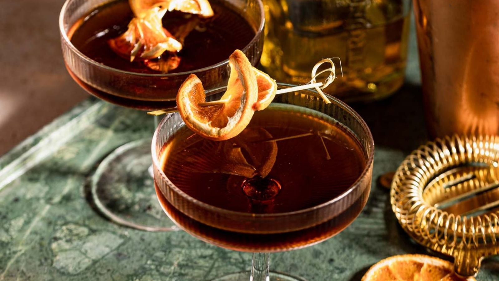 Two coupe glasses filled with dark cocktails, garnished with dried orange slices, rest on a green marble surface.