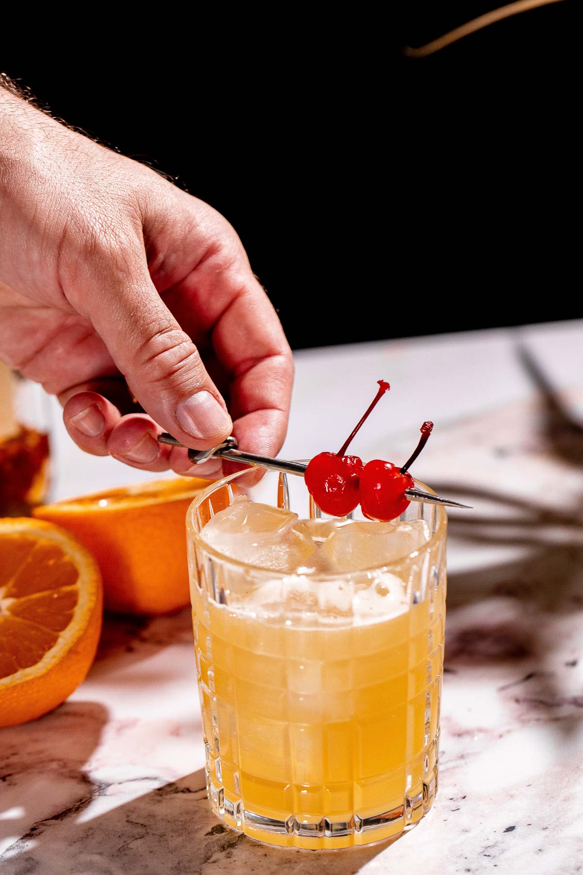 A hand garnishes a glass of amaretto stone sour with a skewer of cherries, while sliced oranges rest on the marble surface nearby.